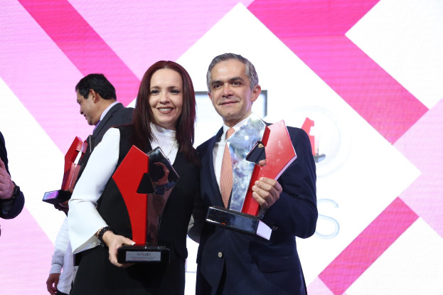 Zwei Personen auf einer Bühne mit Trophäen stehend und strahlend, mit einem Bildschirm im Hintergrund.