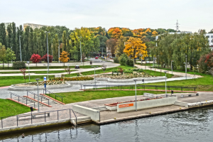 Ein Fluss fließt durch einen grünen Park mit Bäumen, Gras und Pflanzen, neben hohen Gebäuden, Kraftfahrzeugen, Straßenlaternen, Schienen, Geländern, Namensschildern, Strommasten und Kabeln unter einem bewölkten Himmel.