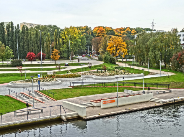 Ein Fluss fließt durch einen grünen Park mit Bäumen, Gras und Pflanzen, neben hohen Gebäuden, Kraftfahrzeugen, Straßenlaternen, Schienen, Geländern, Namensschildern, Strommasten und Kabeln unter einem bewölkten Himmel.