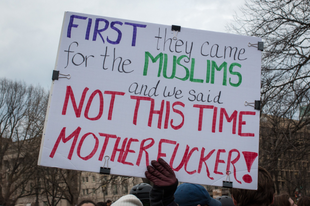 Gruppe von Menschen mit Möppen und Handschuhen, die ein Schild mit der Aufschrift "Zuerst kamen sie wegen der Muslime und wir sagten nicht dieses Mal, verdammte Scheiße" gegen den Hintergrund von Bäumen, Gebäuden und einem klaren blauen Himmel halten.