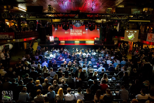 Eine große Menschenmenge sitzt auf Stühlen vor einer Bühne mit Bildschirmen und Bannern, die von Scheinwerfern beleuchtet wird und von Geländern umgeben ist, mit einem Wasserzeichen in der linken unteren Ecke, das eine Atmosphäre der Aufregung und Vorfreude für ein Online-Marketing-Rockstars-Event schafft.