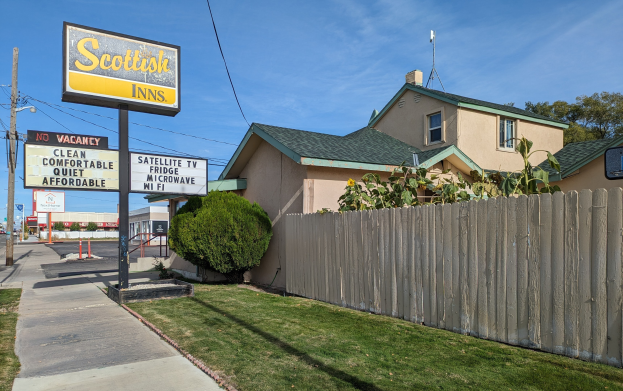 Ein schottisches Motel in San Francisco, Kalifornien, mit einem Haus mit Dach und Fenstern, einem Holzzaun, Pflanzen, Gras, Bäumen, Schildern, Strommasten mit Drähten, einer Straßenlaterne, Fahrzeugen auf der Straße und einem bewölkten Himmel.