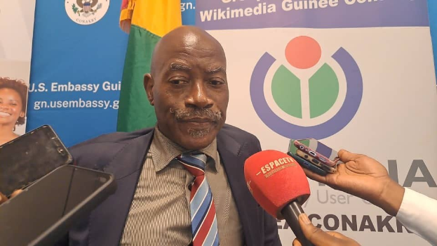 Ein Mann in Anzug und Krawatte spricht mit Reportern, während im Vordergrund eine Hand mit einem Mikrofon und einem Gerät zu sehen ist. Im Hintergrund sind Banner, Flaggen und eine Frau zu erkennen.