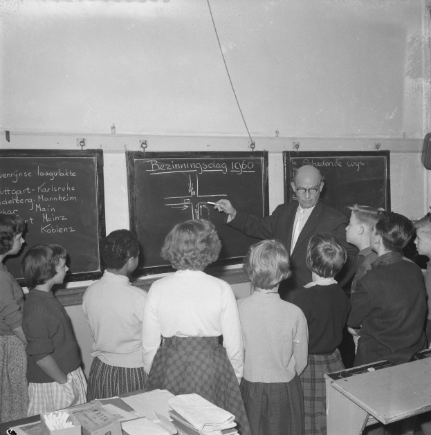 Schwarzes und weißes Foto eines Lehrers, der vor einer Tafel mit einem Marker steht und sitzende Kinder an einem Tisch mit Büchern und Materialien unterrichtet, während auf der rechten Seite ein wandmontierter Lautsprecher zu sehen ist.