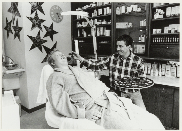 Ein Mann lächelt in einem Zahnarztstuhl, während ein anderer Mann neben ihm steht und eine Platte hält, mit einem Waschbecken, Sternen, einer Uhr und Regalen voller Flaschen im Hintergrund.