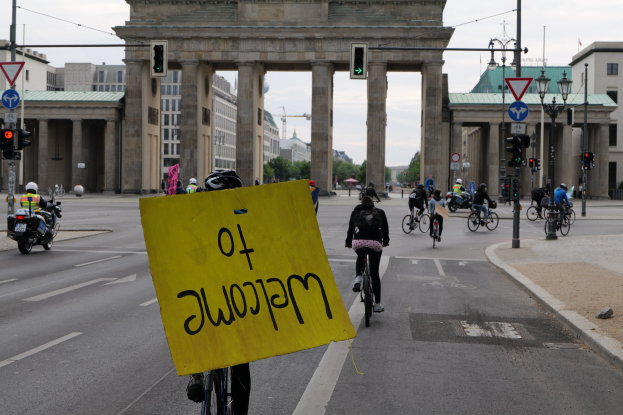 Eine Gruppe von Menschen, die Fahrräder vor dem Brandenburger Tor in Berlin fahren, einer hält ein gelbes Schild mit Text, mit Laternenmasten, Verkehrszeichen, Gebäuden, Bäumen und einem klaren Himmel im Hintergrund.