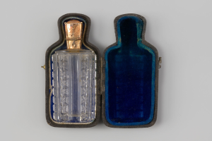 Eine gläserne Parfümflasche mit zylindrischer Form und schmalem Hals, die in einem tiefblauen Etui mit goldener Verzierung auf einer weißen Fläche steht.