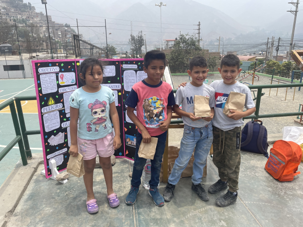 Eine Gruppe von Kindern h├Ąlt Papiert├Ąten in der Hand und steht auf einem Basketballfeld mit T├Ąten und Gegenst├Ąnden auf dem Boden, ein Schild mit Text links daneben und ein Hintergrund aus Z├Ąunung, Pfosten, Stra├čenlaternen, B├Ąumen, Geb├Ąuden, H├Ąngen und Himmel.