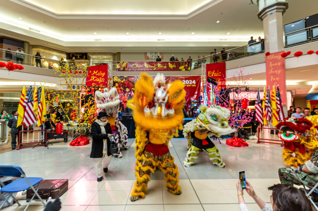 Ein lebendiges chinesisches Neujahrsfest in einem Einkaufszentrum mit tanzenden Menschen in Kostümen, anderen auf Stühlen sitzend und Handys haltend, mit Flaggen, Bannern und dekorativen Elementen im Hintergrund.