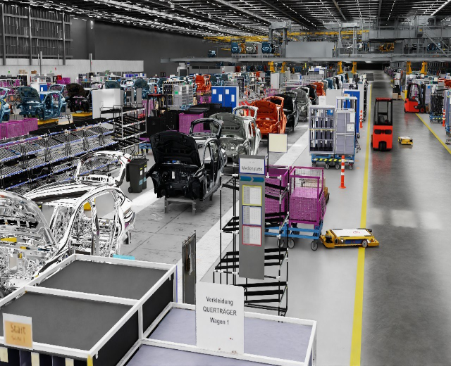 Ein großes Automobilwerk mit mehreren Autos auf der Fertigungsstraße, Metallstrukturen an den Seiten, einer Wand im Hintergrund und an der Decke angebrachten Leuchten, wahrscheinlich eine Volkswagen-Anlage in Deutschland.