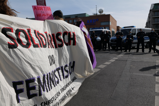 Eine Gruppe von Menschen marschiert mit einer 'Solidarität und Feminismus'-Schlagzeile die Straße entlang, mit parkenden Fahrzeugen, Gebäuden, einer Schüsselantenne und einem klaren blauen Himmel im Hintergrund.