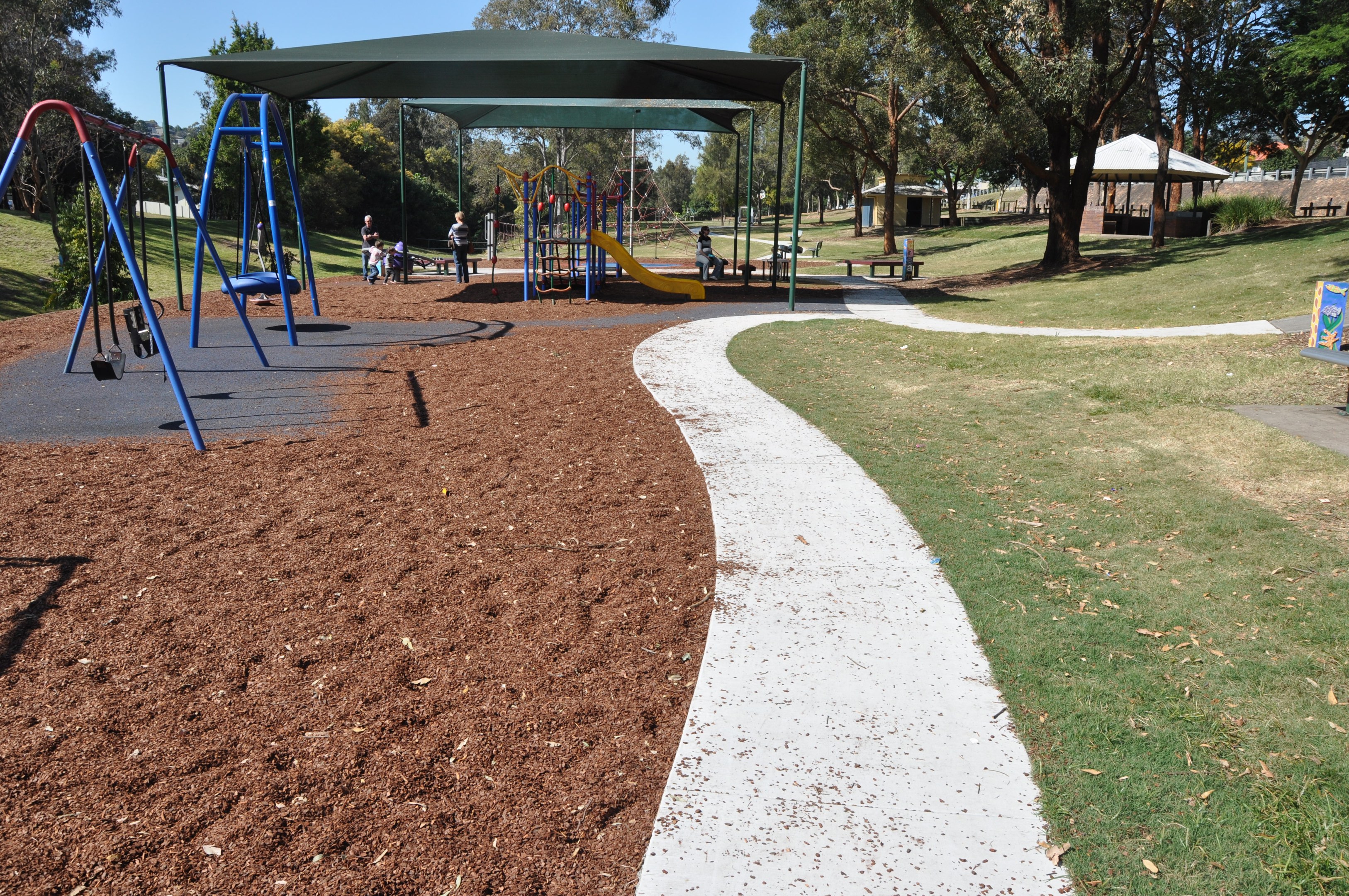 Ein Park mit einem Spielplatz mit Schaukeln und Rutschen, umgeben von grünem Gras, Bäumen, Bänken, Schuppen im Hintergrund und einem klaren blauen Himmel.
