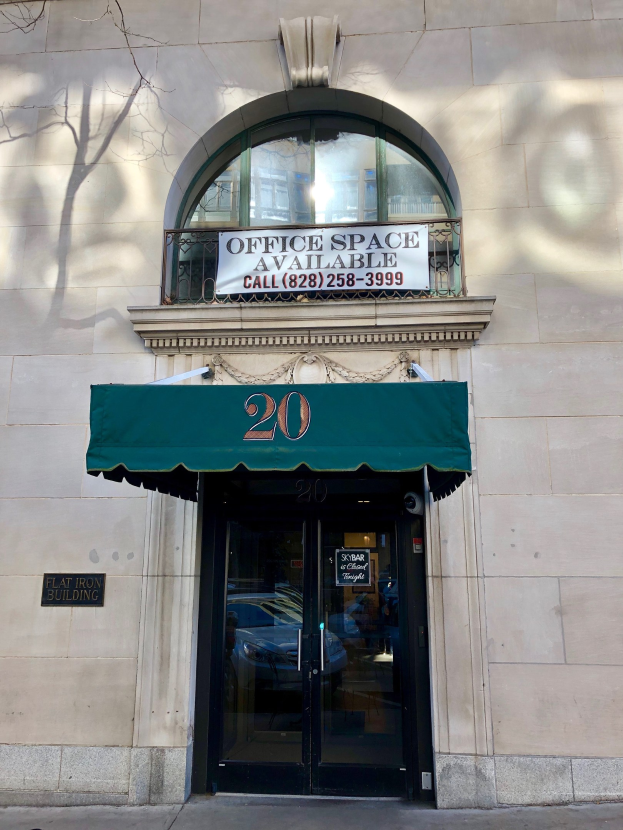 Ein Gebäude mit einer Glastür, einer grünen Markise, einem Schild mit der Aufschrift 'Büroräume zu vermieten' und einem Geländer sowie einem Baum auf der linken Seite.