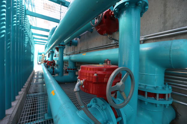 Eine große Gruppe von blauen Rohren und Ventilen in einem Gebäude, mit einem roten Ventil in der Mitte, Metallstangen links und einer Wand im Hintergrund, die wahrscheinlich den Bau einer Gasleitung zeigt.