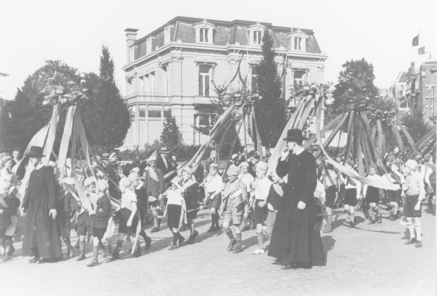 Schwarzes und weißes Bild einer Gruppe von Menschen, die vor einem Gebäude die Straße entlanggehen, einige halten Fahnen, tragen Hüte, mit Bäumen und Gebäuden im Hintergrund und einem Maibaum.