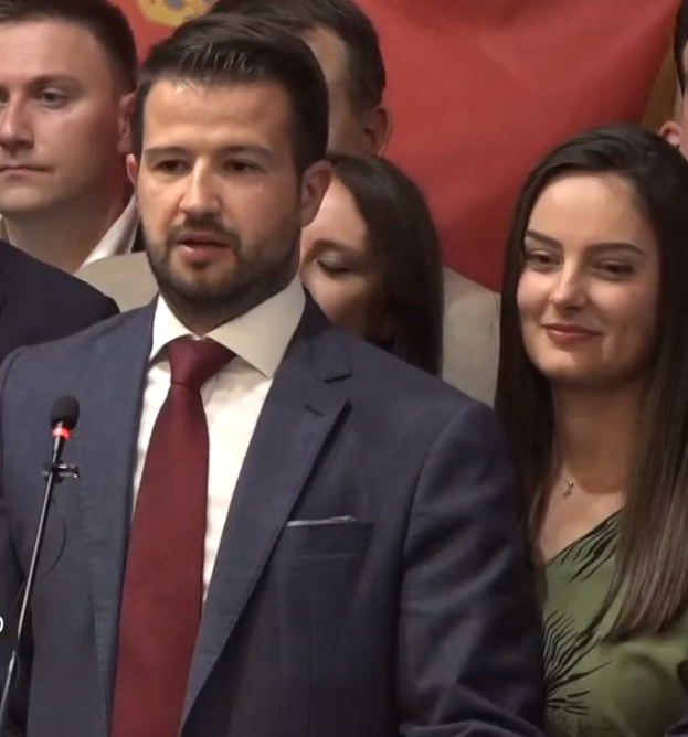 Ein Mann in Anzug und Krawatte spricht in ein Mikrofon an einem Podium, mit einem Banner dahinter und einer aufmerksamen Menge davor.