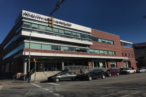 Ein großes Gebäude, das als das New Yorker Gesundheitszentrum für Obdachlose identifiziert wurde, mit Autos davor, Straßenlaternen, Straßenschildern, Menschen auf dem Gehweg, Geländern, Treppen, Bäumen und einem klaren blauen Himmel.