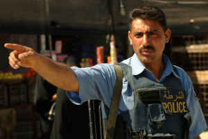 Ein Polizist in Uniform zeigt mit einer Waffe in der Hand auf ein Geschäft mit unscharfen Menschen und Gegenständen im Hintergrund.