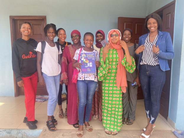 Eine Gruppe von Frauen, die Bücher halten und lächeln, vor einem Schulgebäude in Nigeria stehend, mit einem Tuch auf dem Boden und Türen im Hintergrund.
