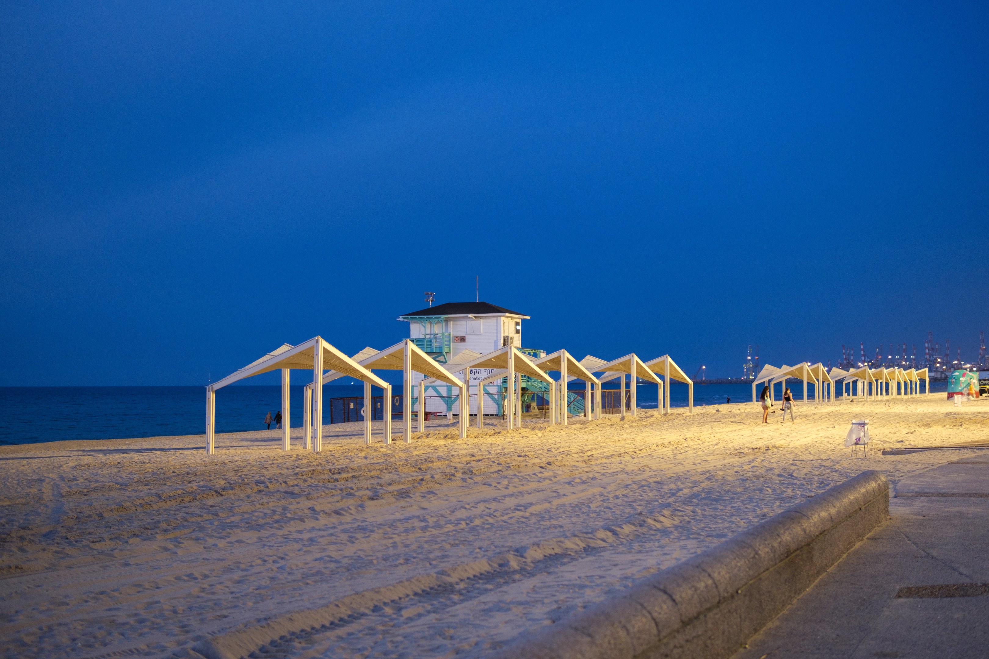 Ein Strand bei Nacht mit einem Rettungsturm, Menschen am Strand, einem Zaun, Zelten, einem Haus, Pfählen, dem Ozean, Lichtern und einem bewölkten Himmel.