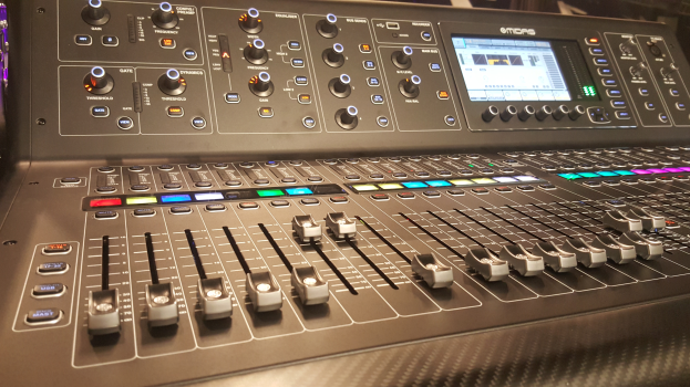 Ein Allen & Heath QU-2400 Digitaler Mischer mit zahlreichen Tasten und einem Bildschirm, der auf einem Tisch steht, mit ein paar unbestimmten Gegenständen im Hintergrund.