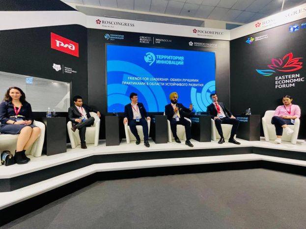 Eine Gruppe von Menschen sitzt auf einer Bühne beim Östlichen Wirtschaftsforum in Moskau, mit Flaschen und anderen Gegenständen auf den Tischen, Bannern mit Text und Logos im Hintergrund und Deckenleuchten.