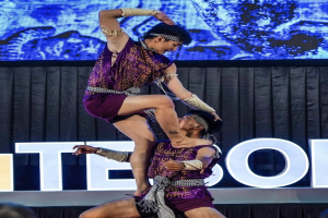 Zwei Performer in violetten Kleidern führen eine akrobatische Hebung auf der Bühne vor einem Publikum aus, mit einem schwarzen Vorhang und Bildschirm im Hintergrund.