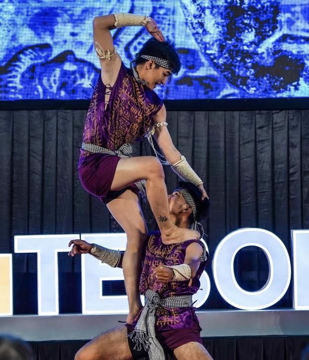 Zwei Performer in violetten Kleidern führen eine akrobatische Hebung auf der Bühne vor einem Publikum aus, mit einem schwarzen Vorhang und Bildschirm im Hintergrund.