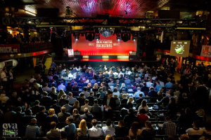 Eine große Menschenmenge sitzt auf Stühlen vor einer Bühne mit Bildschirmen und Bannern, die von Scheinwerfern beleuchtet wird und von Geländern umgeben ist, und erwartet gespannt den Beginn eines Online-Marketing-Rockstars-Events, mit einem Wasserzeichen in der linken unteren Ecke.