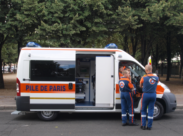 Ein Rettungswagen steht mit drei Personen davor auf der Straße geparkt, mit Bäumen, Gebäuden und anderen Fahrzeugen im Hintergrund.