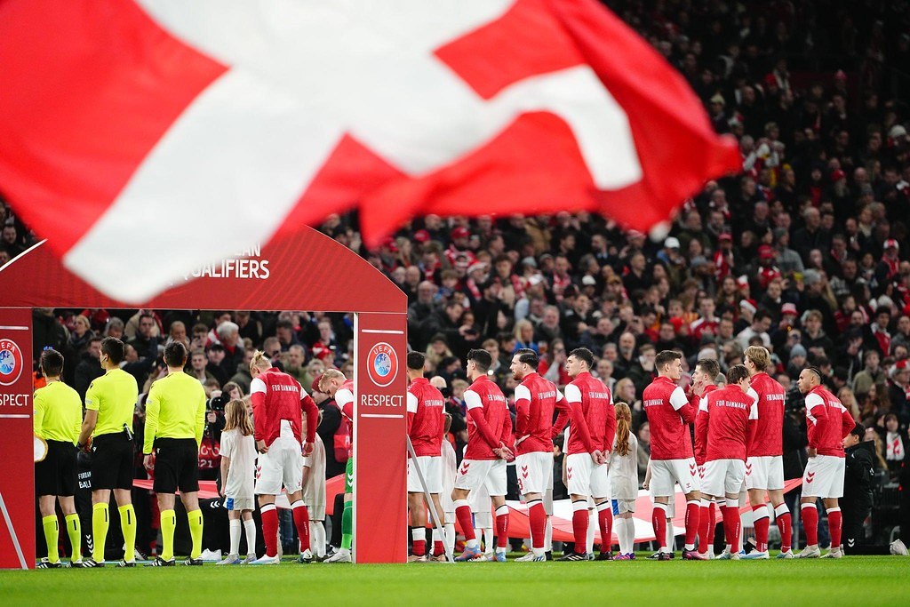 Gruppe von Menschen auf einem Fußballfeld mit einer roten und weißen Flagge im Vordergrund, einem Bogen mit der Aufschrift "Bayern München vs Bayern München Wetten & Vorschau" im Hintergrund und einer großen Menge im Stadion.