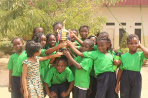 Gruppe von Kindern mit einem Pokal in einem Schulhof mit Bäumen, Pflanzen und einem Gebäude im Hintergrund.