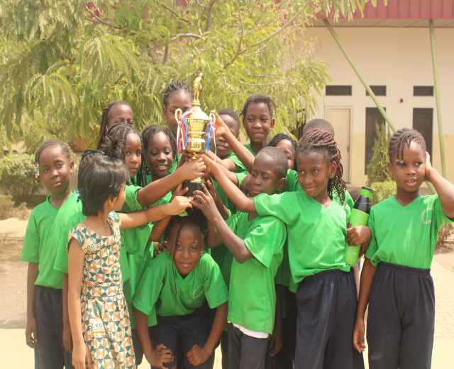Gruppe von Kindern mit einem Pokal in einem Schulhof mit Bäumen, Pflanzen und einem Gebäude im Hintergrund.