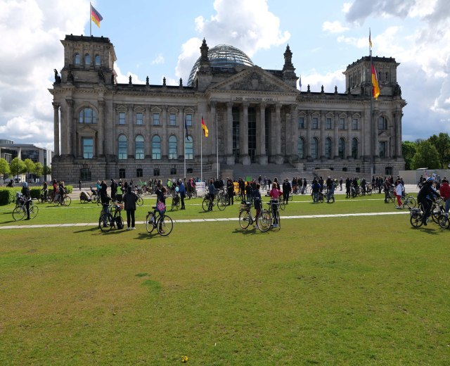 Menschen auf Fahrrädern vor dem Reichstaggebäude in Berlin, Deutschland, mit Bäumen, Pflanzen und einem bewölkten Himmel.