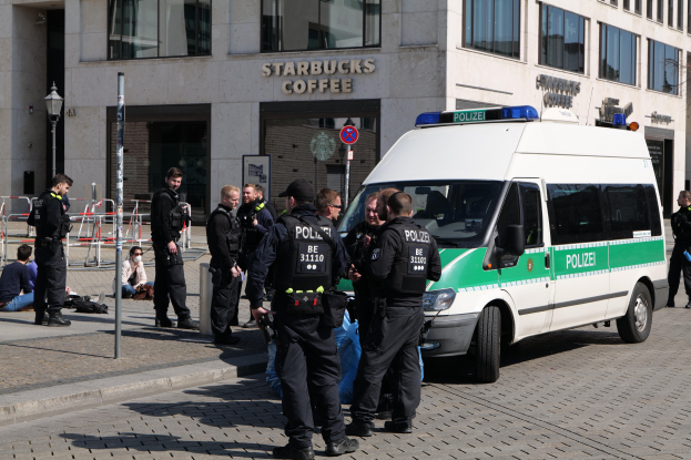 Eine Gruppe von Polizisten steht vor einem Starbucks-Café mit einem Van auf der rechten Seite und einigen Menschen auf der linken Seite, während im Hintergrund ein Gebäude mit Fenstern, ein Schild, ein Laternenmast und ein Zaun zu sehen ist.