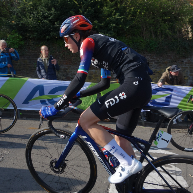 Profi-Radsportlerin Katie Archibald trägt Helm, Schutzbrille, Handschuhe und Schuhe und fährt ein Fahrrad auf einer Straße neben einer Menschenmenge, mit einem Banner, einer Wand, Bäumen und einem Gebäude im Hintergrund.