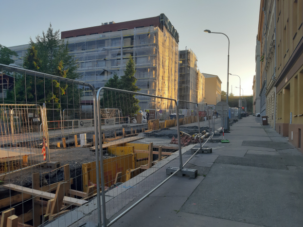 Eine Stadtstraße mit einer Baustelle in der Mitte, umgeben von Gebäuden, Straßenlaternen, Autos, Absperrpoller, Bäumen und einem klaren blauen Himmel.