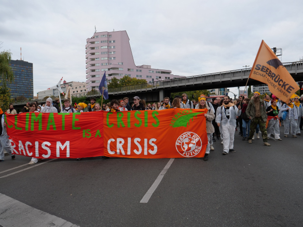 Gruppe von Menschen mit einer "Klima-Krise ist eine Krise"-Plakette, die eine baumbestandene Straße mit Gebäuden und einer Brücke im Hintergrund unter einem bewölkten Himmel entlanggeht.