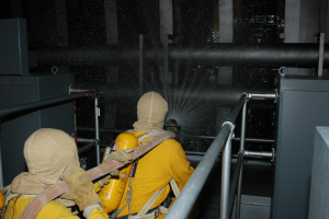 Zwei Feuerwehrleute in gelben Uniformen und Helmen sprühen Wasser aus einem Schlauch auf ein Metallgeländer, mit Metallstangen und anderen Gegenständen im Hintergrund und Text am unteren Bildrand.