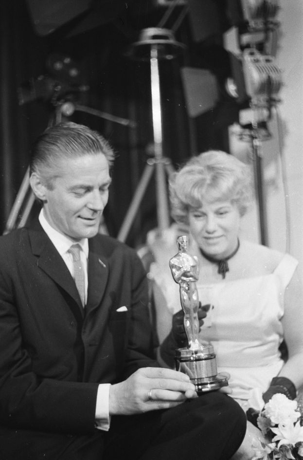 Ein Schwarz-Wei├č-Bild eines formell gekleideten Mannes mit einer Oscar-Statue und einer Frau mit einem Blumenstrau├čus, die gemeinsam sitzen und von Hintergrundbeleuchtung umgeben sind.