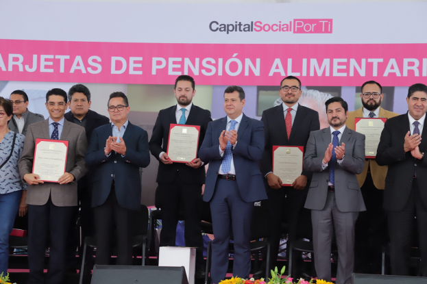 Eine Gruppe von Menschen auf einer Bühne, die klatschen und Zertifikate halten, mit Stühlen in einem Halbkreis darum herum und einem Banner mit der Aufschrift "Capital Social for Ti" im Hintergrund.