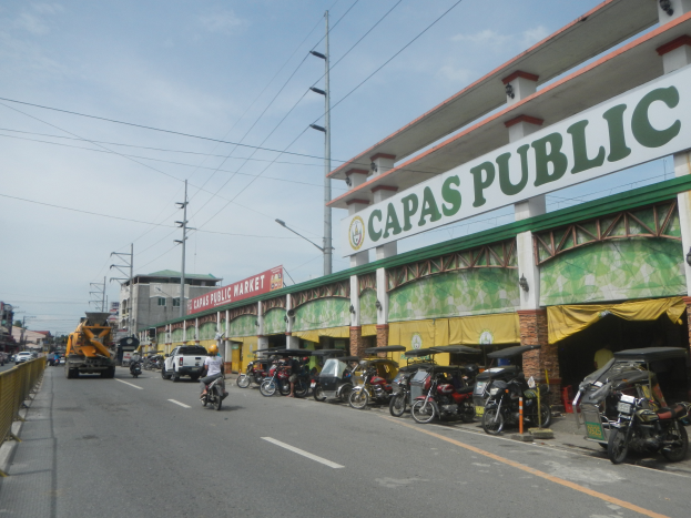 Eine belebte Stadtstraße mit Fahrzeugen, einem Geländer auf der linken Seite, Gebäuden, Polen, Laternen, Schildern und einigen Menschen unter einem bewölkten Himmel, mit einem Gebäude auf der Seite, das ein Schild trägt, auf dem 'Capas Public' steht.