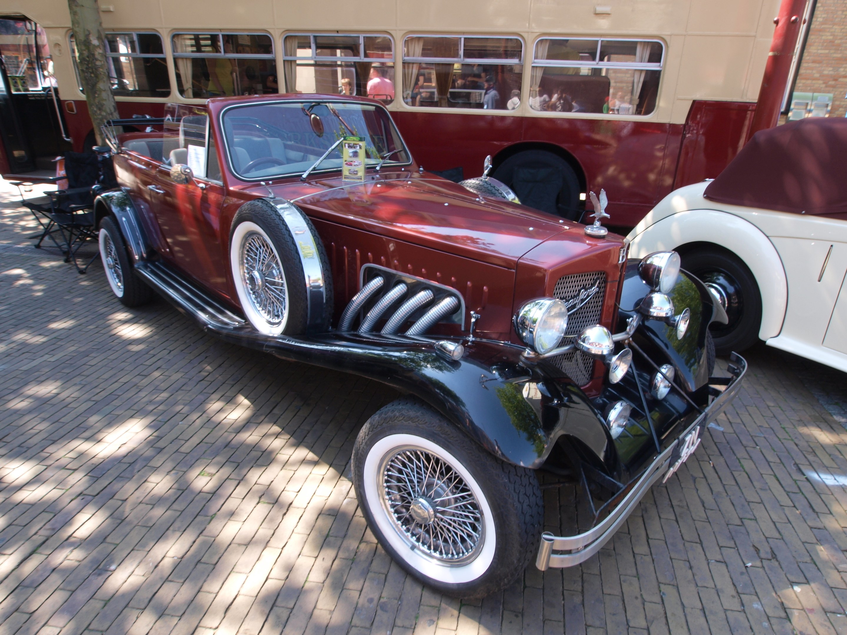 Ein Oldtimer neben einem Doppeldeckerbus auf einem Kopfsteinpflasterweg geparkt, mit Stühlen und einem Baum im Vordergrund, einem Gebäude im Hintergrund und Menschen im Bus zu sehen.