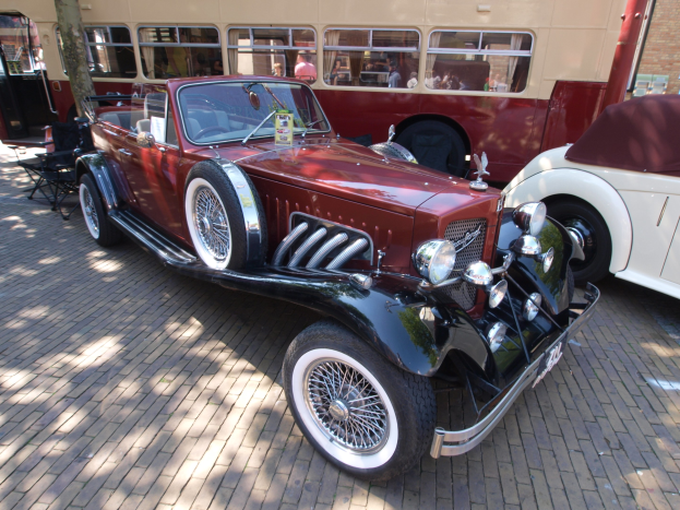 Ein Oldtimer neben einem Doppeldeckerbus auf einem Kopfsteinpflasterweg geparkt, mit Stühlen und einem Baum im Vordergrund, einem Gebäude im Hintergrund und Menschen im Bus zu sehen.