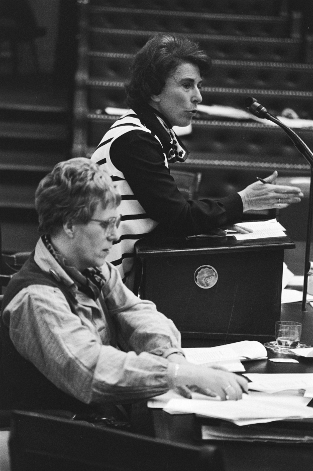 Schwarz-weiß-Foto einer Frau, die an einem Tisch im Europäischen Parlament in Straßburg im Mai 1981 sitzt und in ein Mikrofon spricht, während sie einen Stift hält und Papiere, ein Glas Wasser und andere Gegenstände auf dem Tisch liegen.