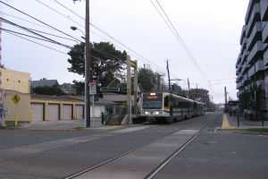 Eine Straßenbahn fährt durch eine Straße mit hohen Gebäuden, Strommasten und einigen Fußgängern, mit Bäumen und einem klaren blauen Himmel im Hintergrund.