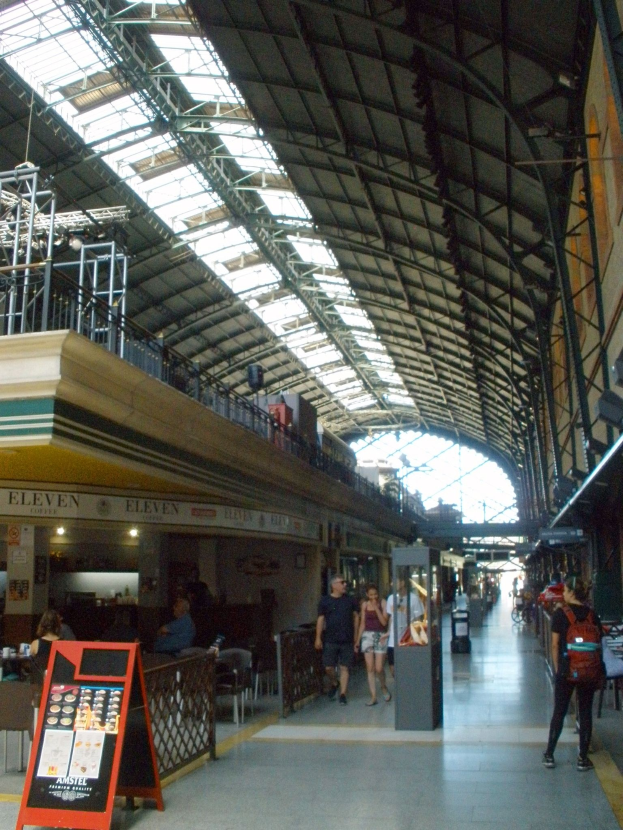 Menschen mit Gepäck durch einen Bahnhof gehen, Abfahrtsanzeige, Sitzplätze und Mülltonne sind zu sehen.