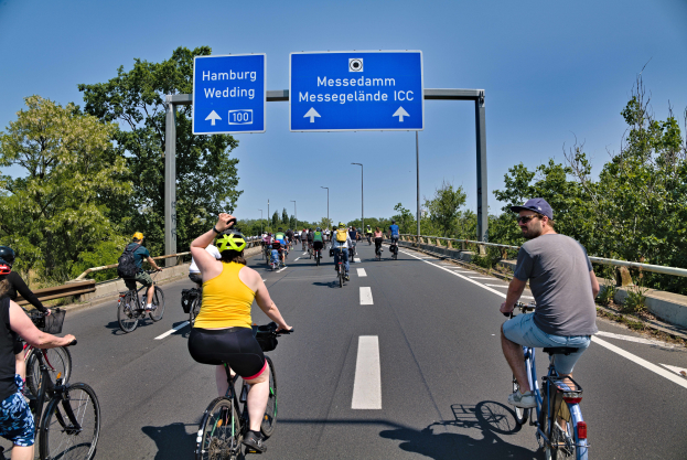 Eine Gruppe von Radfahrern in Helmen fährt auf einer Straße mit einer Begrenzung auf einer Seite und Bäumen auf der anderen, unter einem klaren blauen Himmel mit Laternen im Hintergrund und einer Texttafel oben.