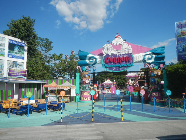 Karussell am Parkeingang mit Straße, eingezäuntem Bereich, Buden, Banner, Bäumen und bewölktem Himmel im Hintergrund.