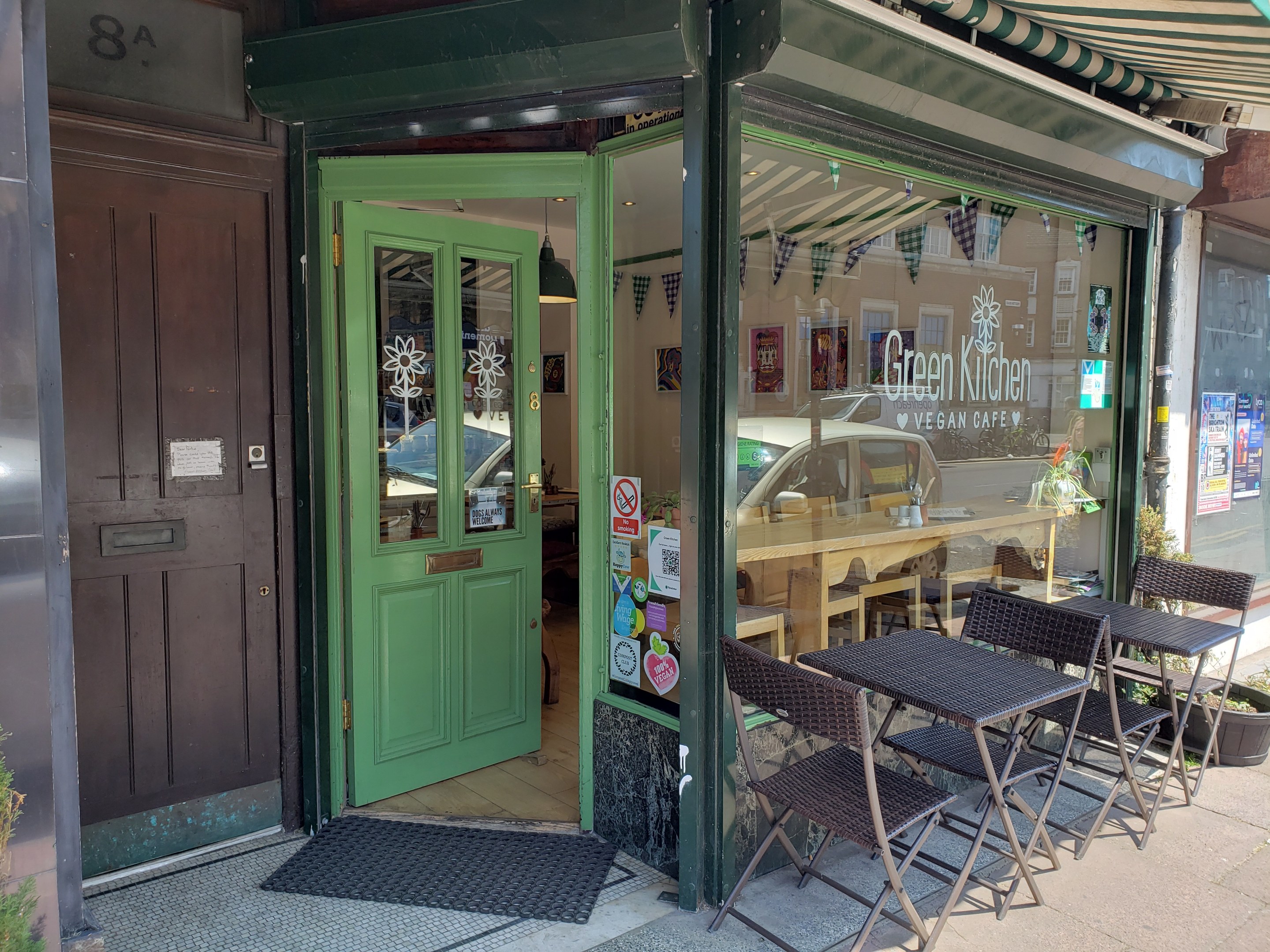 Außensitzbereich des Green Kitchen Vegan Cafe mit Tischen, Stühlen, Pflanzen, einer Matte, einer Glastür und Postern an der Glaswand; drinnen sind Tische, Stühle und eine Hängelampe zu sehen.